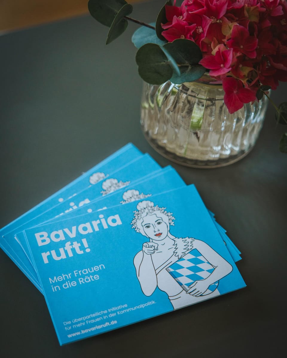 Blaue Flyer der überparteilichen Initiative Bavaria ruft mit dem Motto Mehr Frauen in die Räte liegen neben einer Vase mit rosa Blüten auf einem Tisch