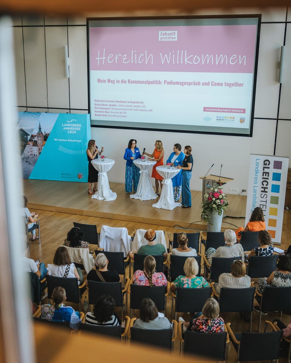 Fünf Frauen stehen beim Podiumsgespräch an weißen Stehtischen auf einer Bühne, im Hintergrund eine Leinwand mit dem Motto Zukunft gestalten, davor das sitzende Publikum