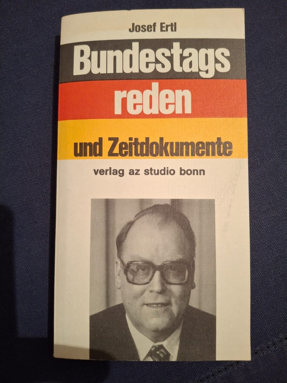 Buchtitel „Josef Ertl, Bundestagsreden und Zeitdokumente“ in Schwarz-Rot-Gold mit einem Schwarz-Weiß-Porträt Josef Ertls mit Brille, das Buch liegt auf dunkelblauem Stoff