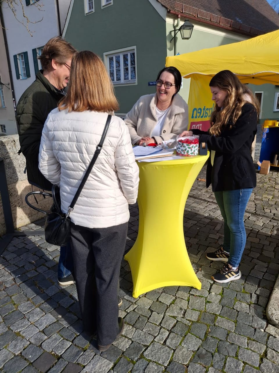 Ulla Schäfer sitzt am gelben Stehtisch mit der Unterschriftenliste und einer Dose roter Bonbons im Gespräch mit zwei Standbesuchern