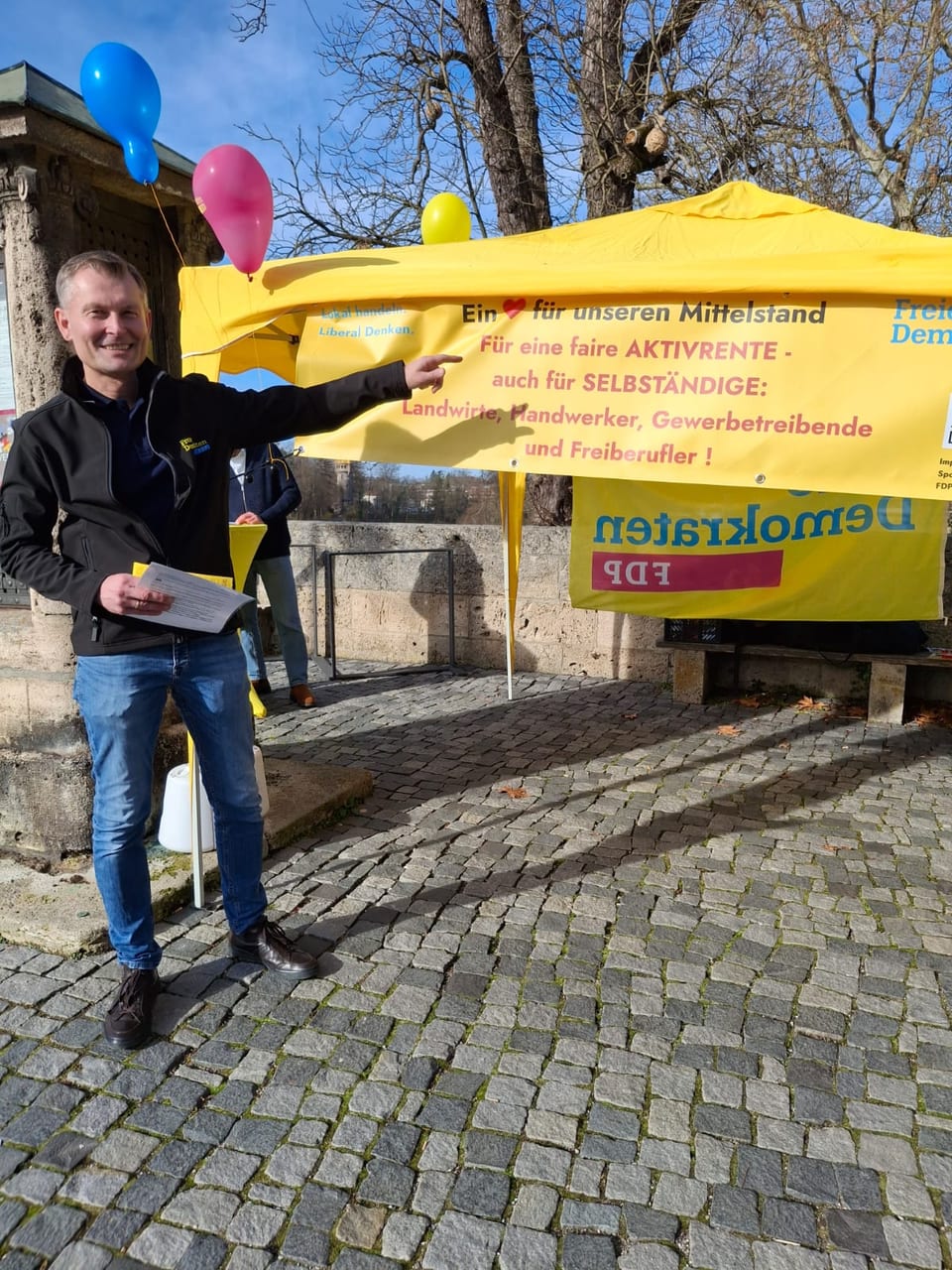 Ein Standmitglied zeigt lächelnd auf das Banner mit der Forderung nach einer fairen Aktivrente auch für Selbständige