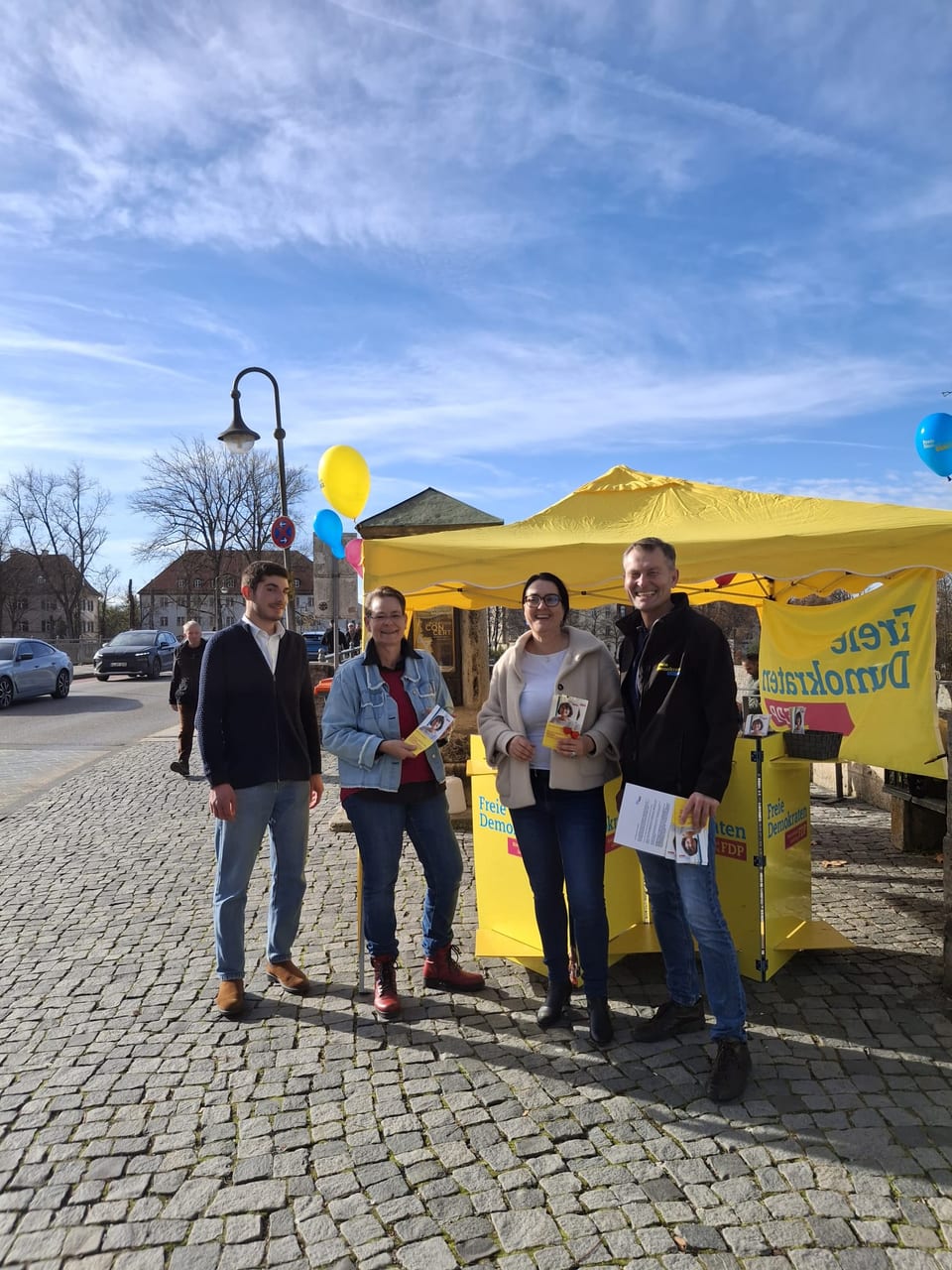 Vier Personen stehen bei Sonnenschein vor dem gelben Infostand der Freien Demokraten an der Karolinenbrücke, dekoriert mit gelben, blauen und pinken Luftballons