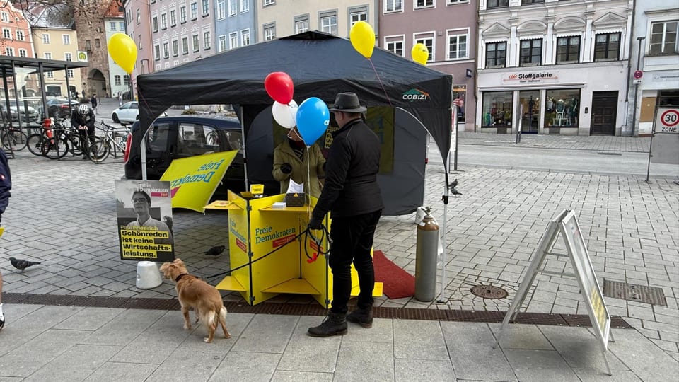 Blick auf den FDP-Infostand mit schwarzem Pavillon und Luftballons auf dem Hauptplatz, im Vordergrund wird ein Ballon mit Gas befüllt, daneben ein Hund an der Leine