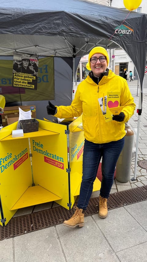 Ulla Schäfer in gelber FDP-Jacke und gelber Mütze am Infostand des Kreisverbands, in der Hand ein Flyer „Aus Liebe zur Freiheit"