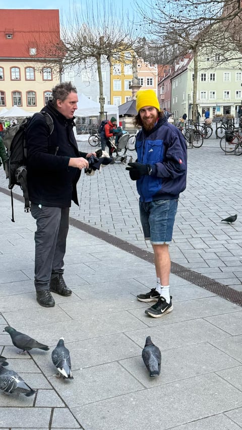 Ein FDP-Standhelfer mit gelber Mütze im Gespräch mit einem Passanten auf dem Hauptplatz, im Vordergrund Tauben