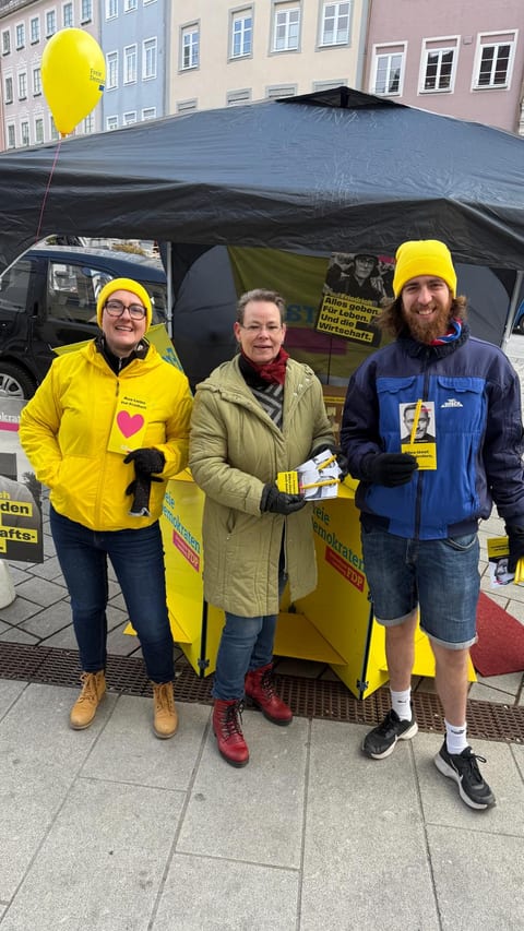 Drei Standhelfer der FDP mit Flyern vor einem Infostand mit schwarzem Pavillon auf dem Landsberger Hauptplatz, im Hintergrund ein Wahlplakat von Paul Friedrich