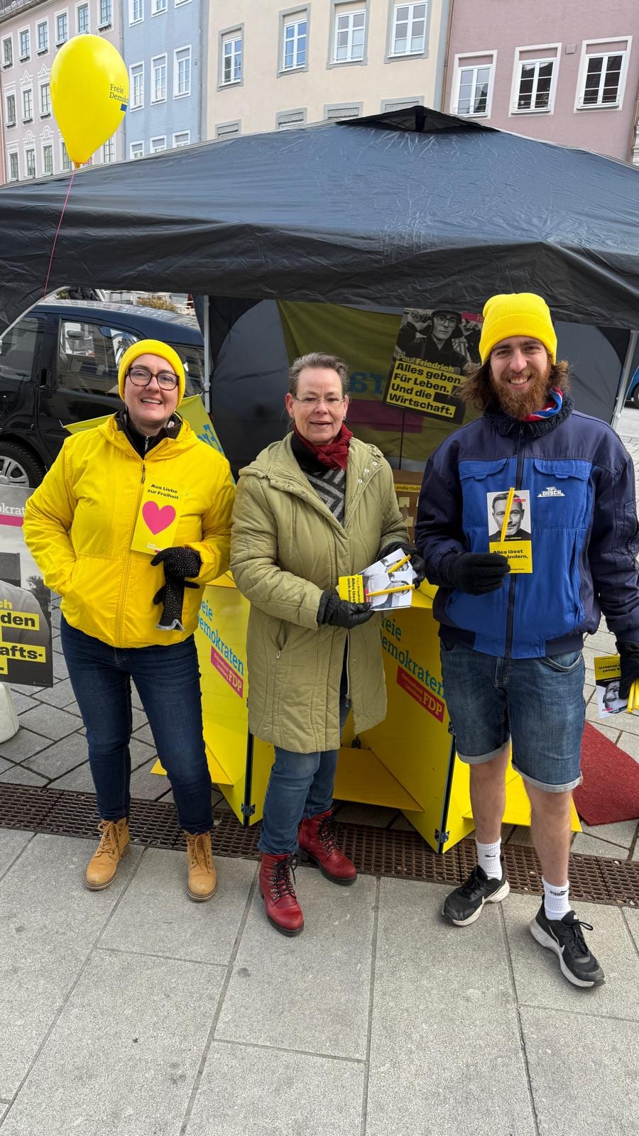 Drei Standhelfer der FDP mit Flyern vor einem Infostand mit schwarzem Pavillon auf dem Landsberger Hauptplatz, im Hintergrund ein Wahlplakat von Paul Friedrich