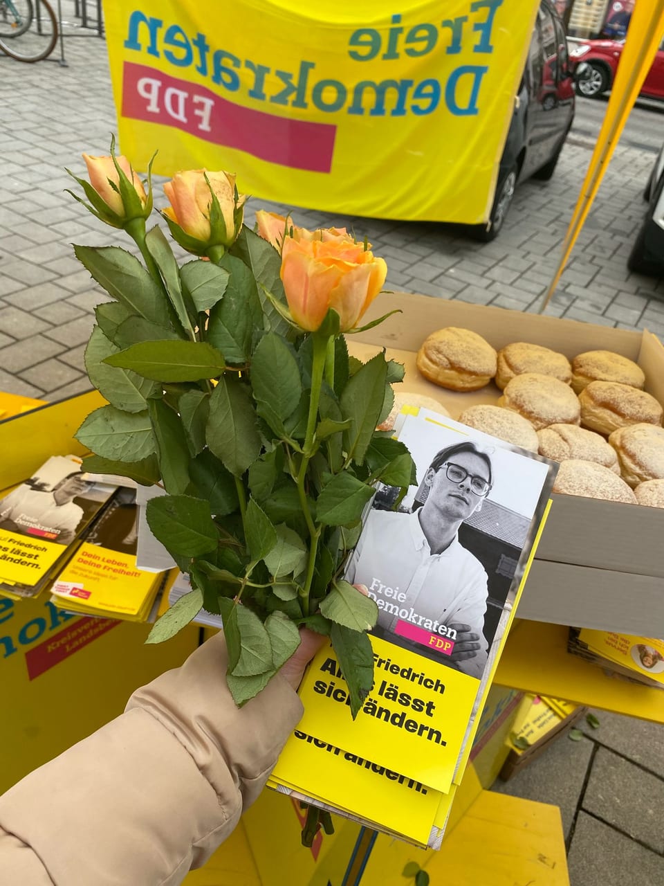 Eine Hand hält einen Strauß gelb-oranger Rosen, daneben FDP-Flyer mit dem Porträt von Paul Friedrich und eine Kiste mit Krapfen