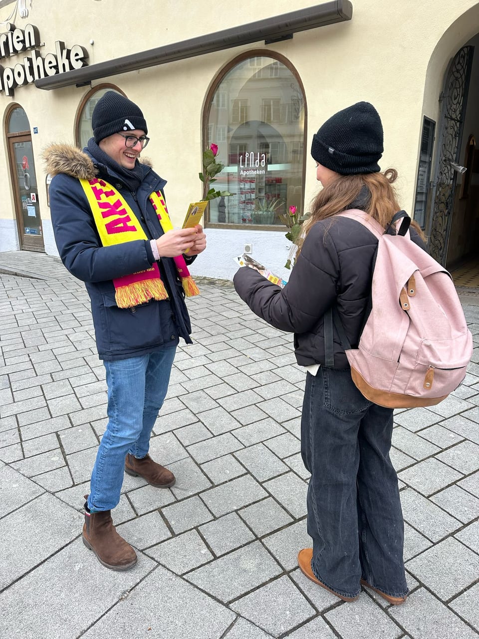Paul Friedrich mit gelb-pinkfarbenem JuLi-Schal überreicht einer jungen Passantin eine Rose und einen Flyer in der Fußgängerzone