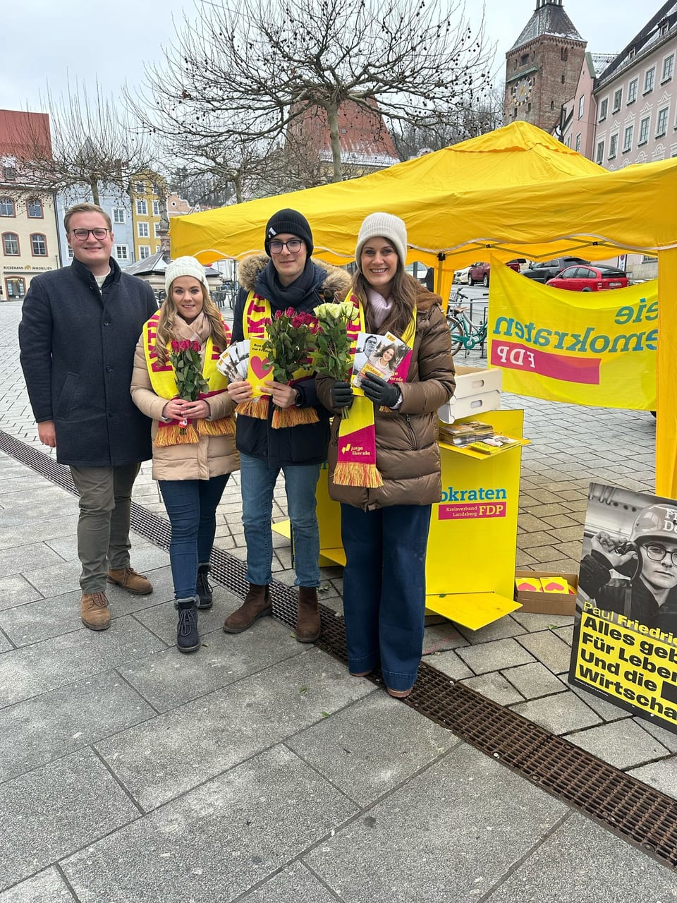 Vier junge Menschen mit Rosen und FDP-Flyern vor einem gelben FDP-Pavillon auf dem Landsberger Hauptplatz, im Hintergrund der Schmalzturm