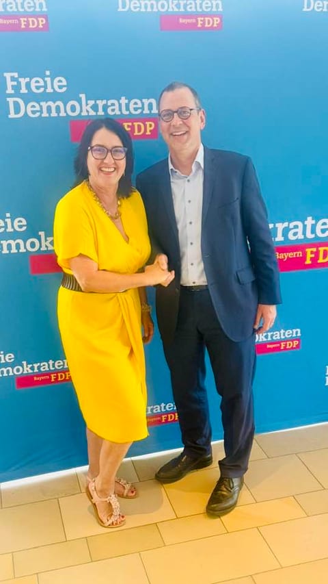 Ulla Schäfer im gelben Kleid gratuliert dem neuen Landesvorsitzenden Dr. Michael Ruoff per Handschlag vor einer blauen Wand mit dem Logo der Freien Demokraten Bayern