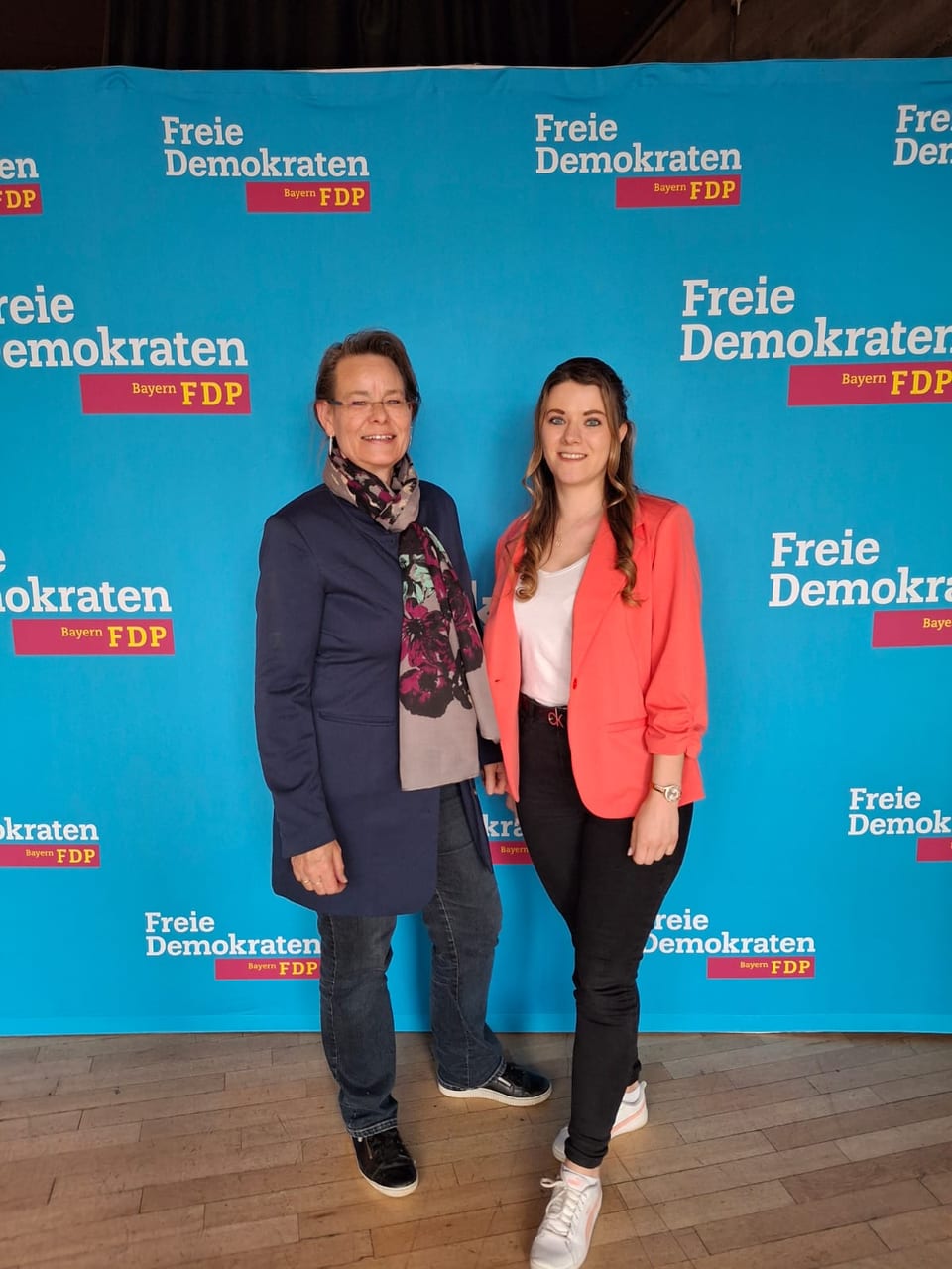 Zwei Delegierte stehen nebeneinander vor einer blauen Fotowand mit dem Logo der Freien Demokraten Bayern, links eine Frau mit Brille und Blazer, rechts eine jüngere Frau mit korallenfarbenem Blazer