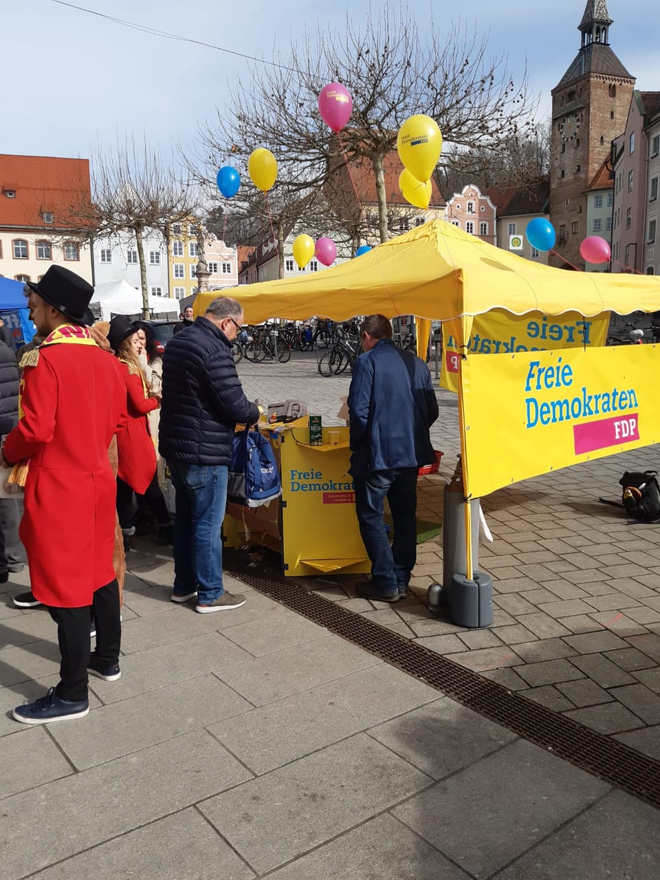 Bürger im Gespräch am gelben FDP-Stand „Freie Demokraten“ auf dem Landsberger Hauptplatz, im Hintergrund der Schmalzturm, links kostümierte Standhelfer