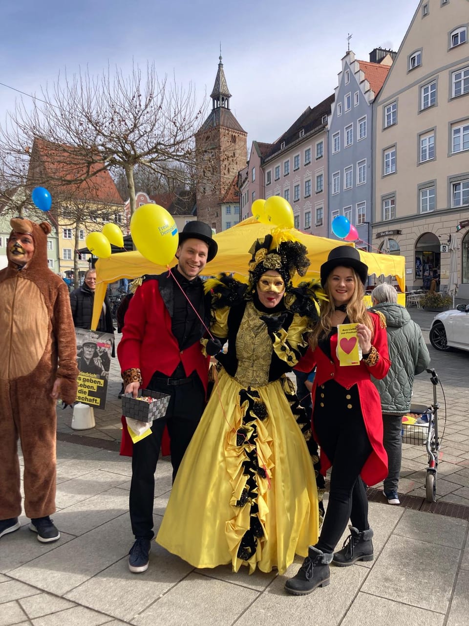 Drei kostümierte Mitglieder des Standteams vor dem gelben FDP-Pavillon: zwei junge Aktive in roten Zirkusdirektor-Kostümen mit Zylinder und in der Mitte Stadt- und Kreisrätin Ulla Schäfer im goldgelben venezianischen Kostüm mit Maske, im Hintergrund der Schmalzturm
