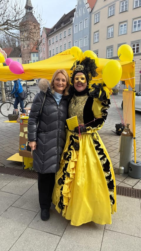 Landsbergs Oberbürgermeisterin Doris Baumgartl im grauen Wintermantel (links) und Stadt- und Kreisrätin Ulla Schäfer im goldgelben venezianischen Kostüm mit Maske vor dem gelben FDP-Stand