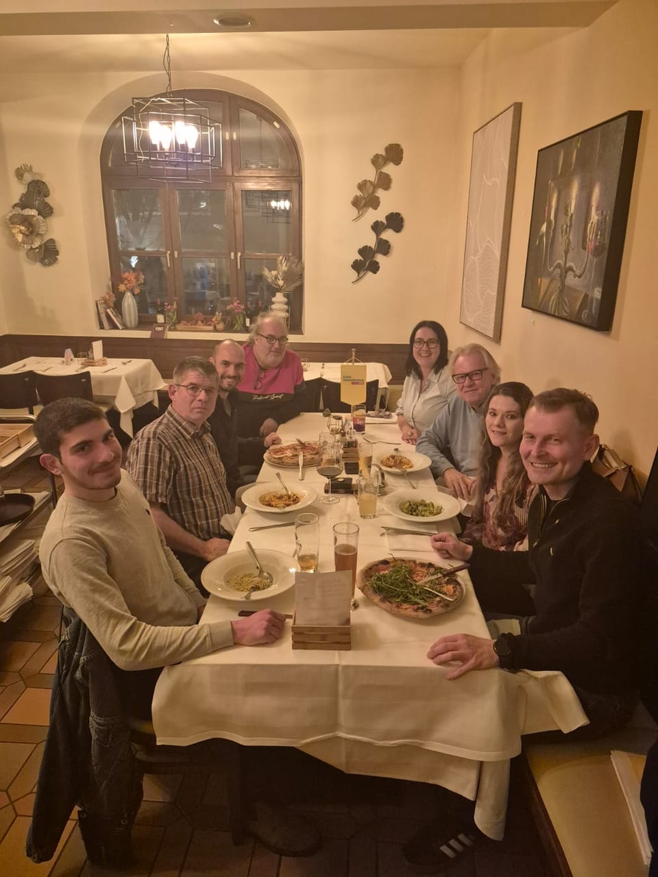 Rund acht Teilnehmerinnen und Teilnehmer sitzen beim Liberalen Stammtisch an einem langen Tisch in der Pizzeria mit Pizza, Pasta und einem gelben Freie-Demokraten-Tischfähnchen