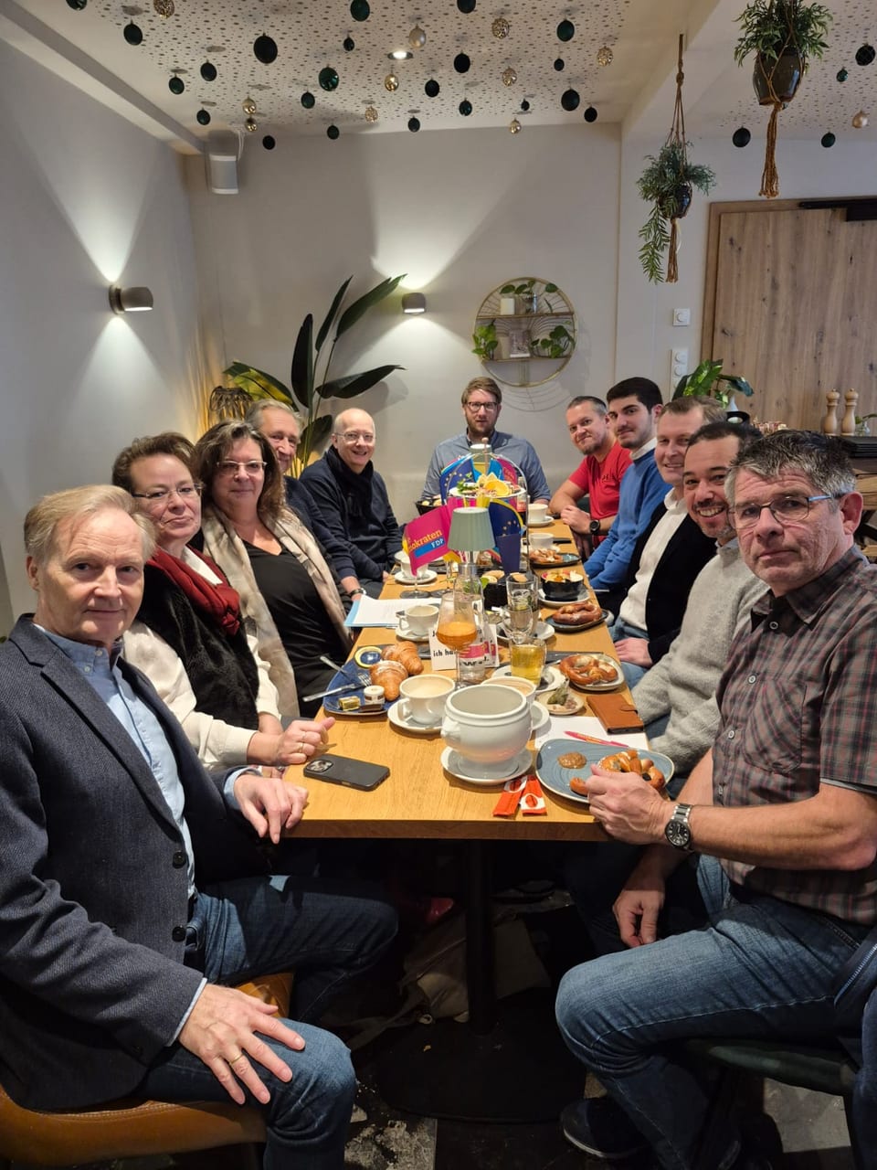 Gruppenbild der Frühstücksrunde an einer langen Tafel in einem gemütlichen Lokal mit Brezen, Croissants und Kaffee sowie Fähnchen der Freien Demokraten und der Europäischen Union