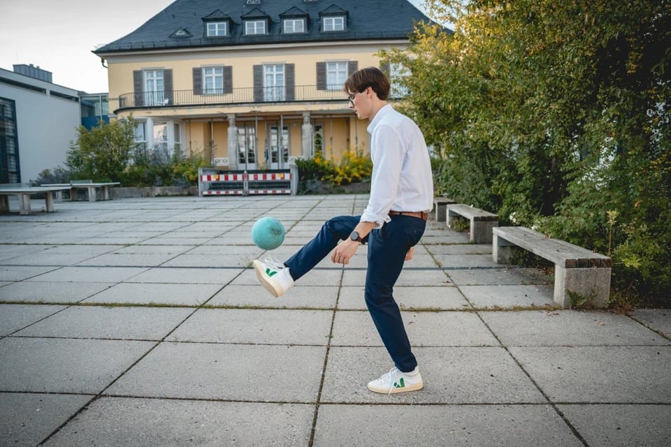 Paul Friedrich in weißem Hemd balanciert eine Weltkugel auf dem Fuß auf einer gepflasterten Terrasse vor einer gelben Villa