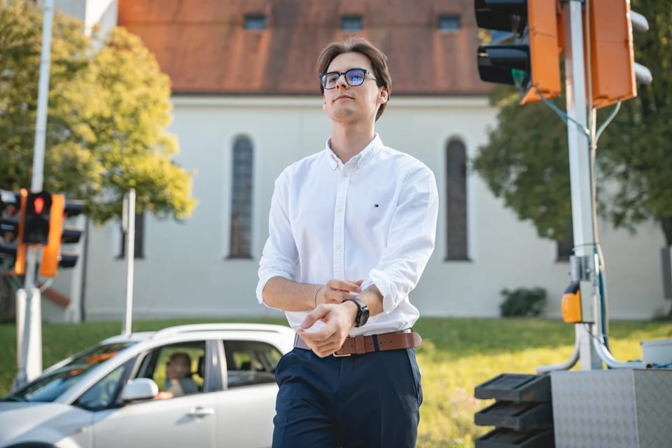 Paul Friedrich in weißem Hemd blickt an einer Ampelkreuzung auf seine Armbanduhr, im Hintergrund eine weiße Kirche