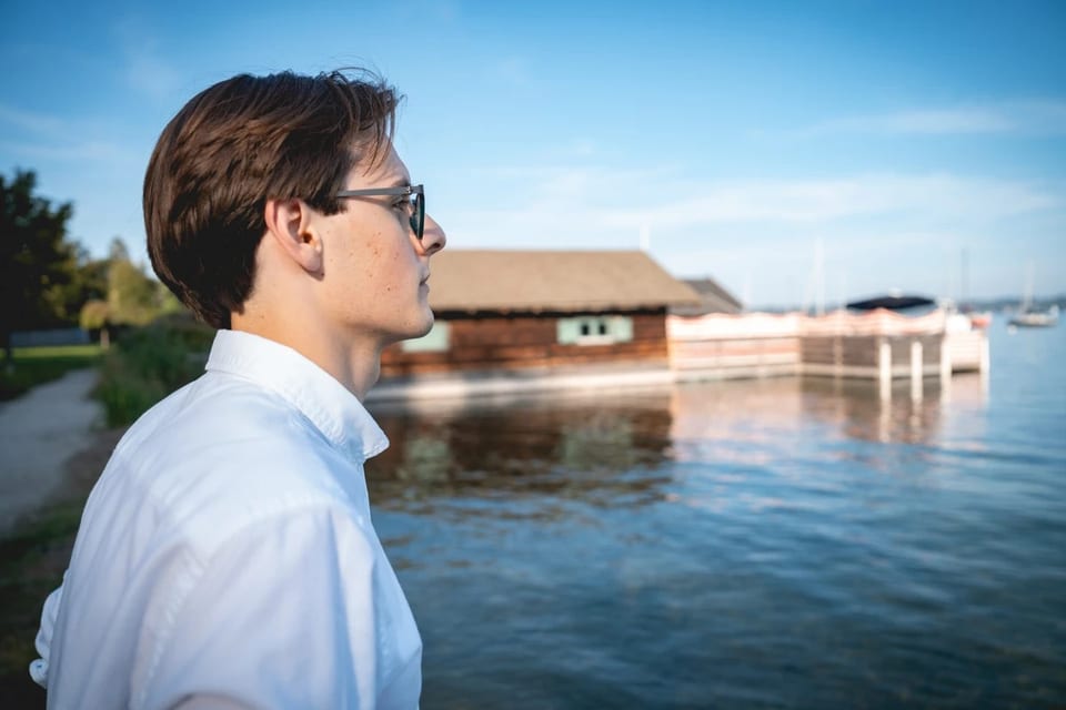 Paul Friedrich im Profil in weißem Hemd blickt über einen See mit Bootshaus und Segelbooten in die Ferne