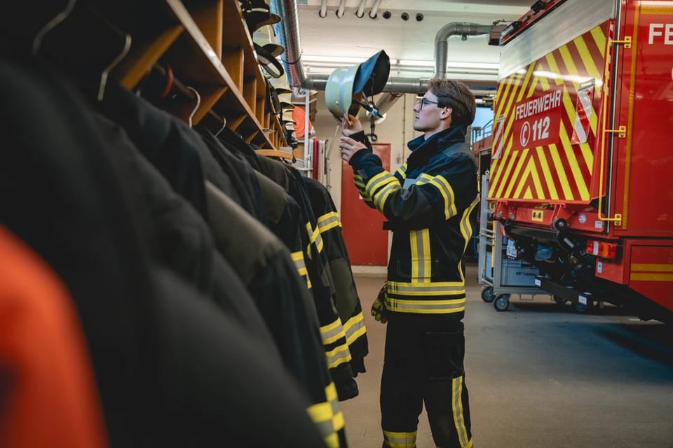 Paul Friedrich in Feuerwehrmontur mit gelben Reflexstreifen prüft in einer Fahrzeughalle einen Helm, daneben ein rotes Feuerwehrfahrzeug mit der Aufschrift Feuerwehr 112