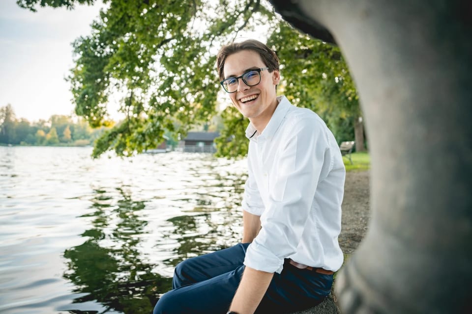 Paul Friedrich sitzt lachend in weißem Hemd an einem Seeufer unter einem Baum, im Hintergrund Wasser und ein Bootshaus