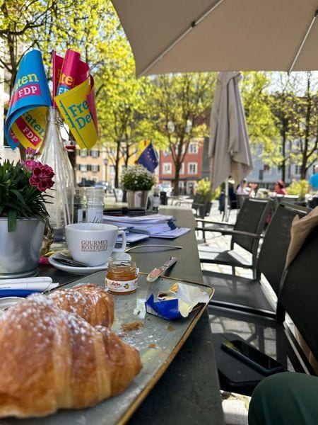 Gedeckter Frühstückstisch im Freien mit Croissants, Kaffee und FDP-Tischfähnchen, im Hintergrund die Landsberger Altstadt