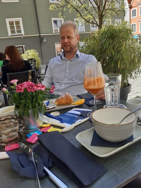 Ein Gast sitzt an einem Frühstückstisch im Freien mit Croissant, Getränk und Informationsmaterial, im Hintergrund Altstadthäuser