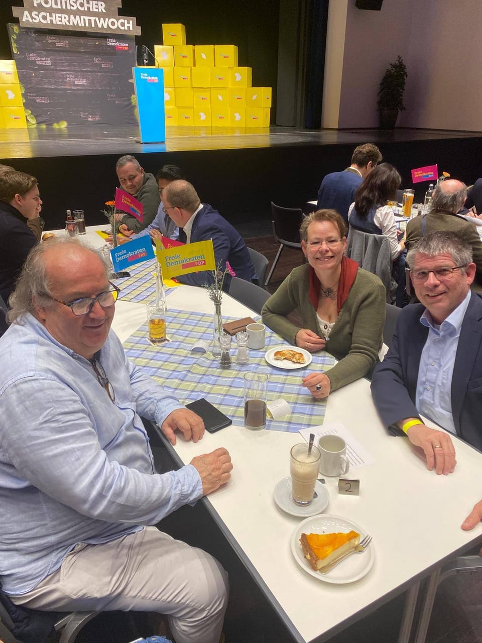 Mitglieder des Kreisverbands Landsberg und des Kreisverbands Miesbach sitzen an einem festlich gedeckten Tisch mit roten FDP-Tischfähnchen, Kaffee und Kuchen, im Hintergrund die Bühne mit der Aufschrift Politischer Aschermittwoch
