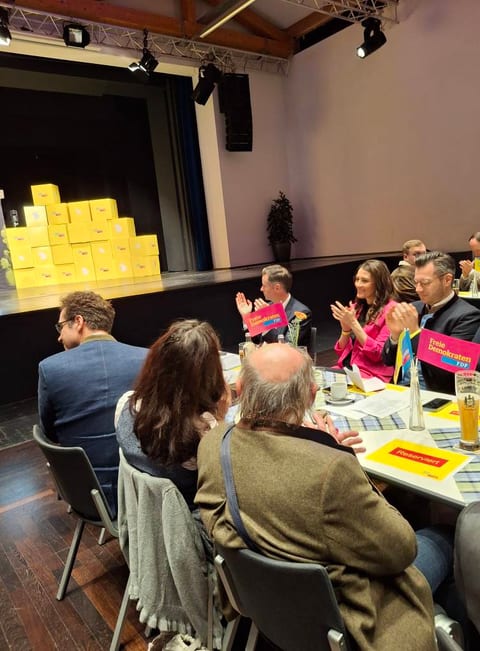 Blick aus dem Publikum auf die vordere Tischreihe beim Politischen Aschermittwoch: Gäste applaudieren, auf dem Tisch stehen rote FDP-Tischfähnchen, im Hintergrund die Bühne mit gestapelten gelben Kartons