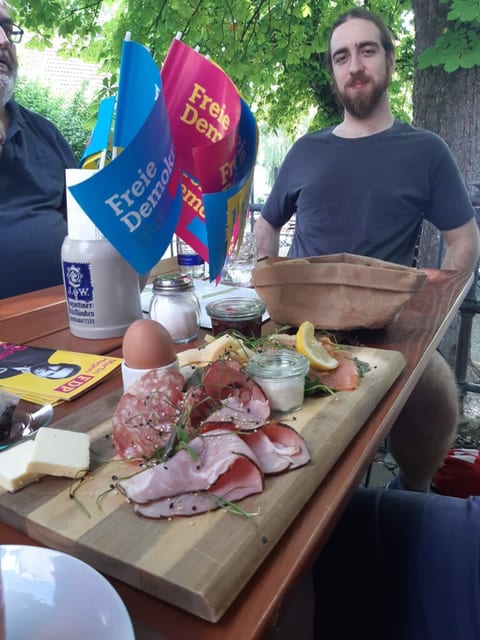 Ein Gast sitzt im Biergarten hinter einem Holzbrett mit Brotzeit aus Wurst, Käse, Ei und Lachs, daneben FDP-Tischfähnchen und ein Bierkrug