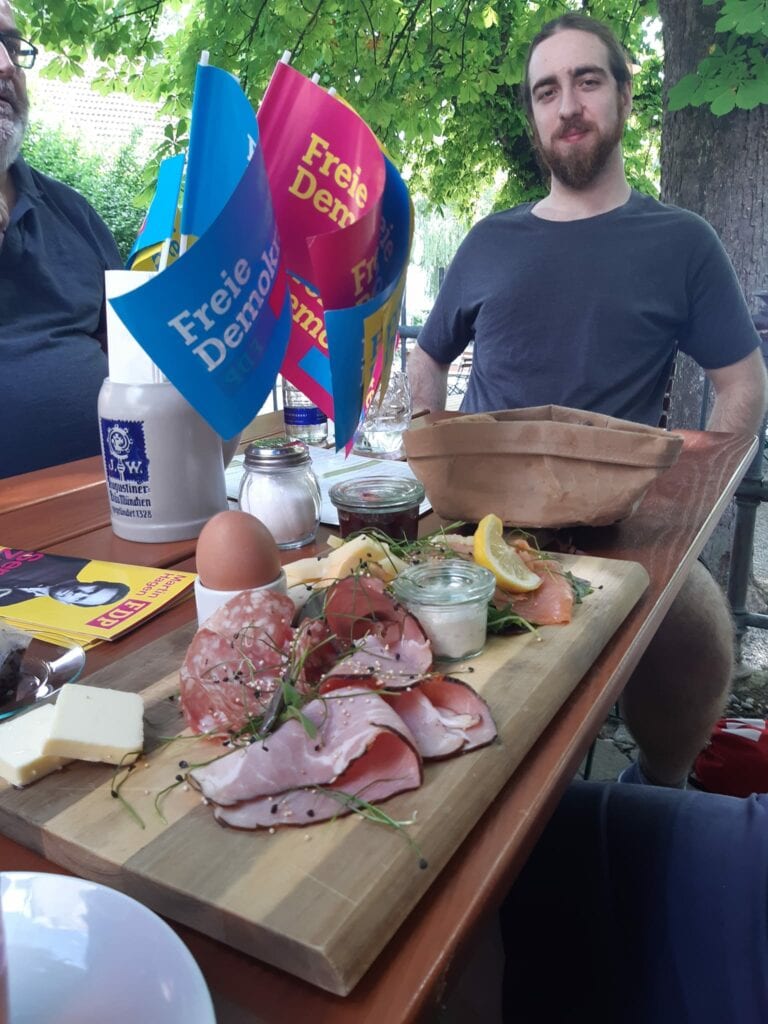 Ein Gast sitzt im Biergarten hinter einem Holzbrett mit Brotzeit aus Wurst, Käse, Ei und Lachs, daneben FDP-Tischfähnchen und ein Bierkrug