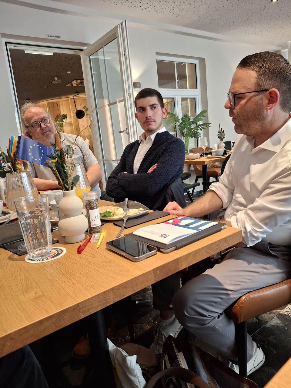 Dr. Constantin Groth im weißen Hemd mit hochgekrempelten Ärmeln spricht am Tisch, ihm gegenüber hört ein jüngerer Mann mit verschränkten Armen zu, links ein weiterer Gast mit Brille; auf dem Tisch eine Europafahne und ein Ukraine-Fähnchen