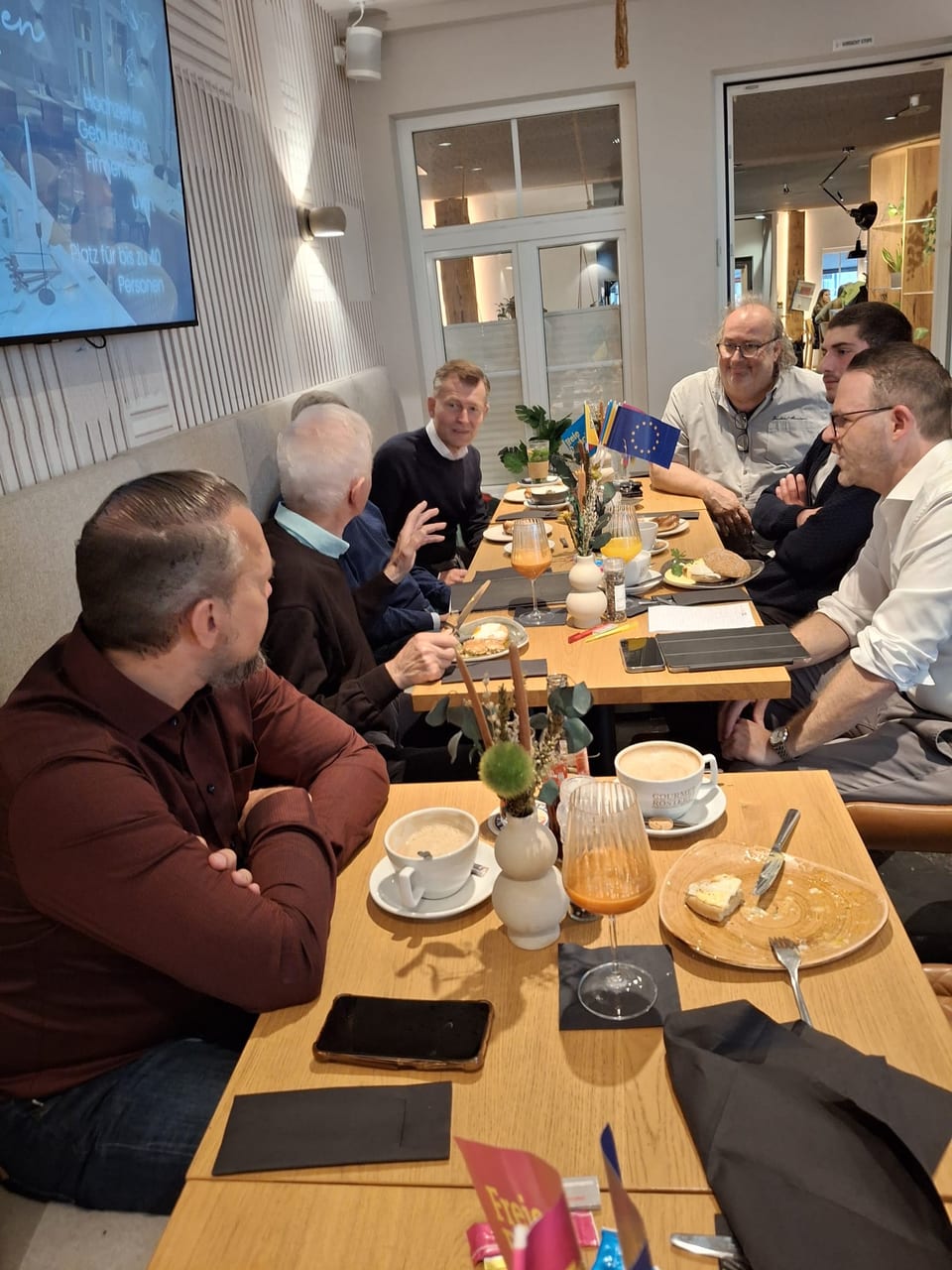 Blick entlang des Frühstückstisches mit Gästen zu beiden Seiten, im Vordergrund ein Mann im dunkelroten Hemd mit verschränkten Armen, dazwischen Kaffeetassen, Orangensaft und FDP-Tischfähnchen