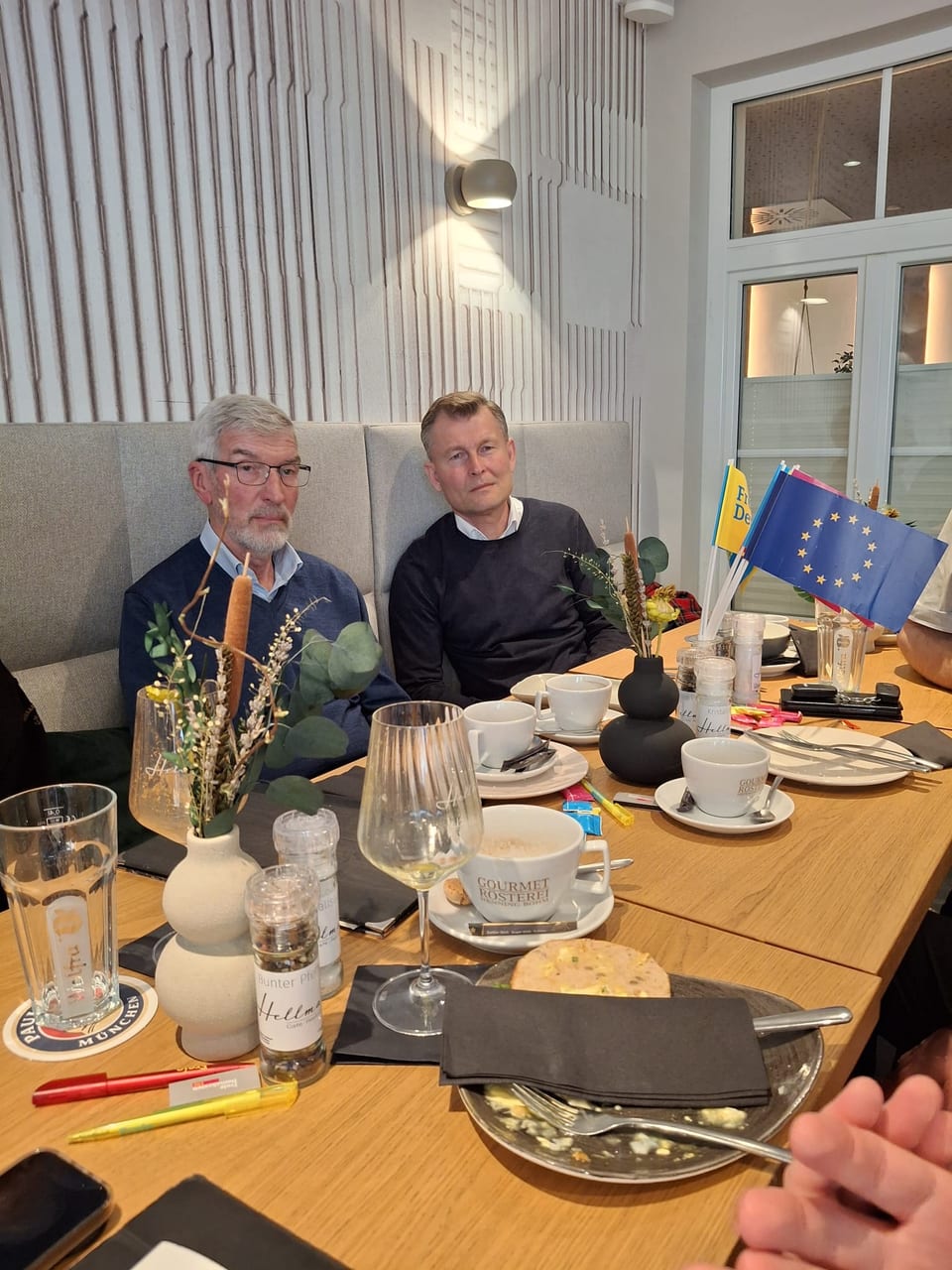 Zwei Gäste sitzen nebeneinander auf einer Sitzbank am Frühstückstisch, auf dem Tisch stehen eine Europafahne und ein kleines Ukraine-Fähnchen in einer Vase sowie Kaffeetassen und Teller