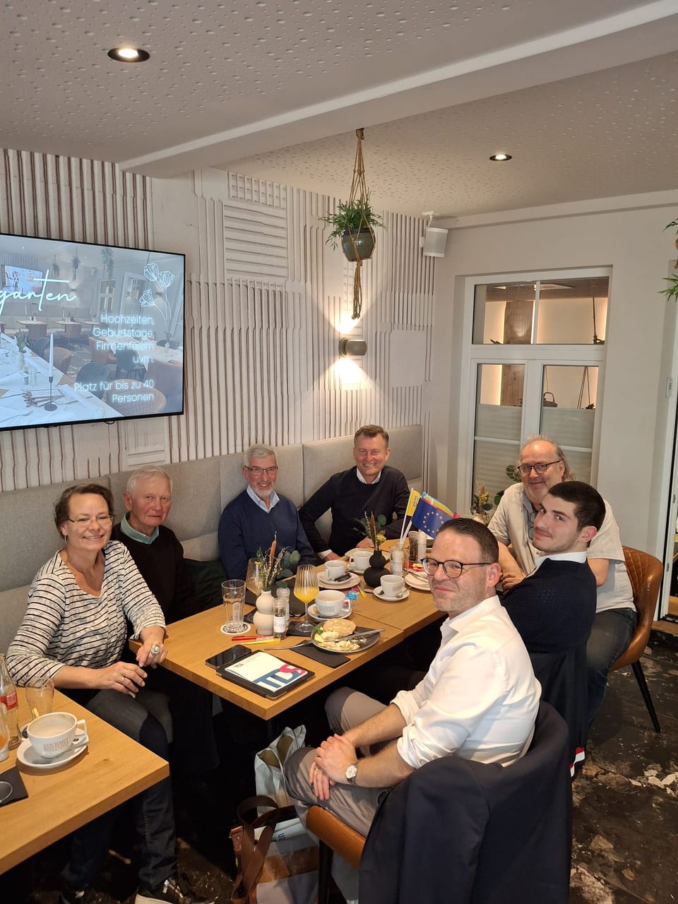 Sieben Gäste sitzen beim Politischen Frühstück um einen Tisch in einem hellen Café, auf dem Tisch eine Europafahne und ein kleines Ukraine-Fähnchen sowie Kaffeetassen und Frühstücksteller