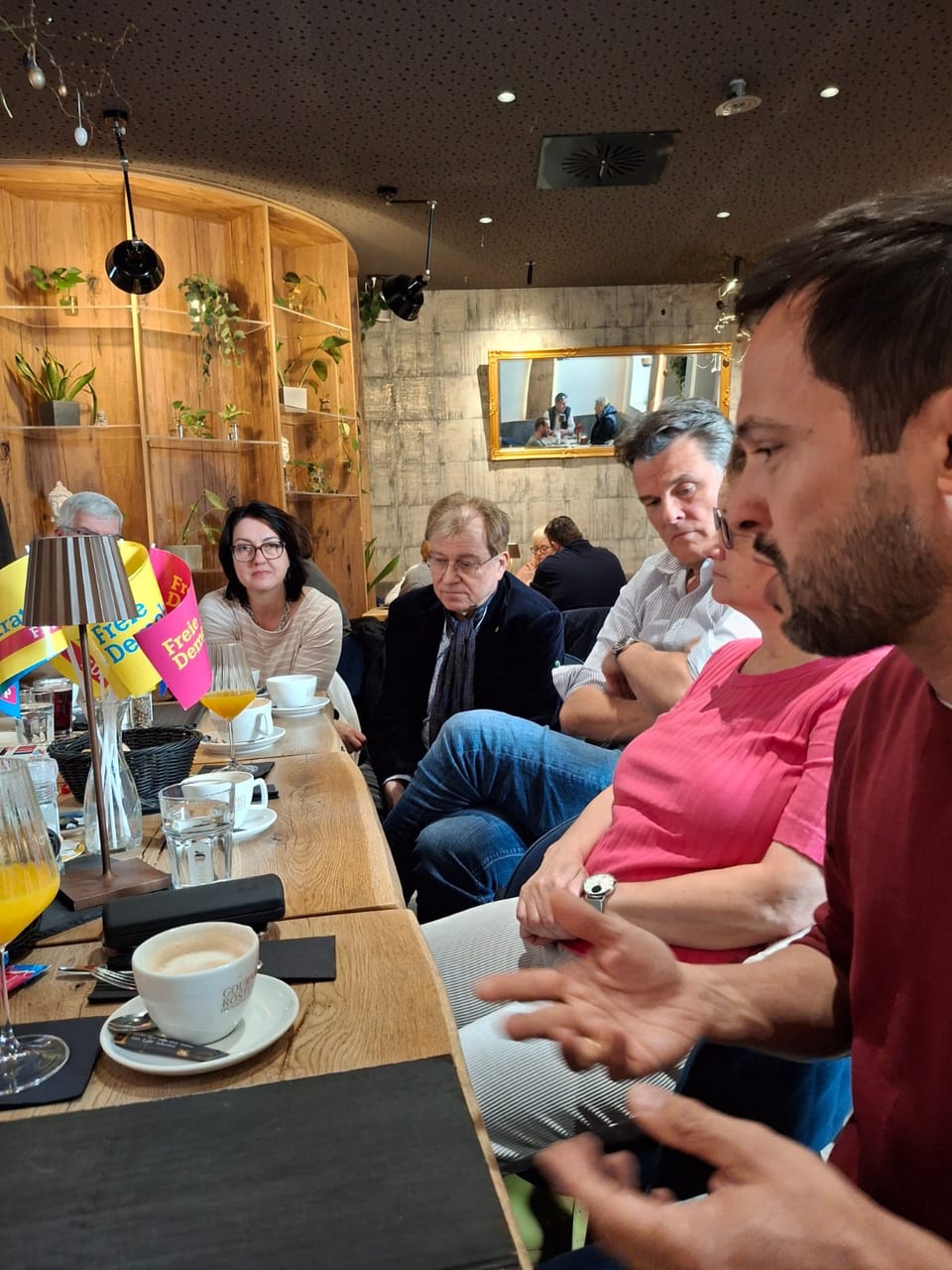 Martin Hagen mit Bart und grauem Jackett spricht am Tisch und gestikuliert mit den Händen, während die Gäste bei Kaffee und Orangensaft aufmerksam zuhören