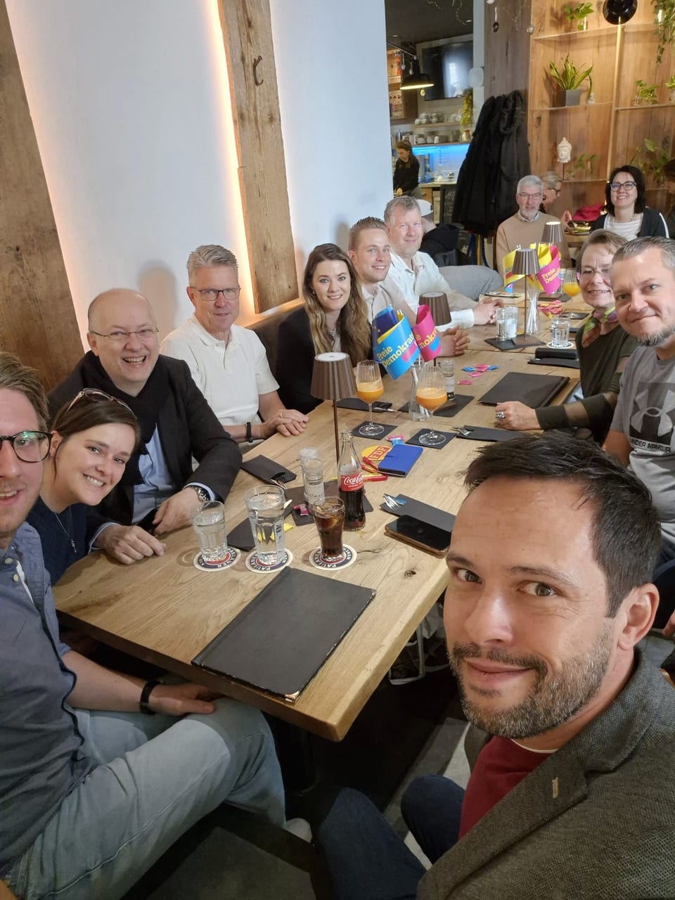 Gruppenselfie der Frühstücksrunde an einem langen Holztisch im Café, auf dem Tisch FDP-Tischfähnchen und Gläser mit Orangensaft, im Vordergrund ein Mann mit Bart